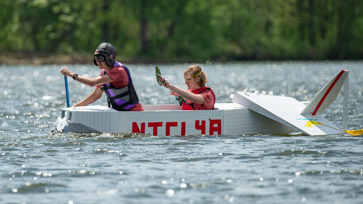 Two people are paddling on the water in a cardboard boat