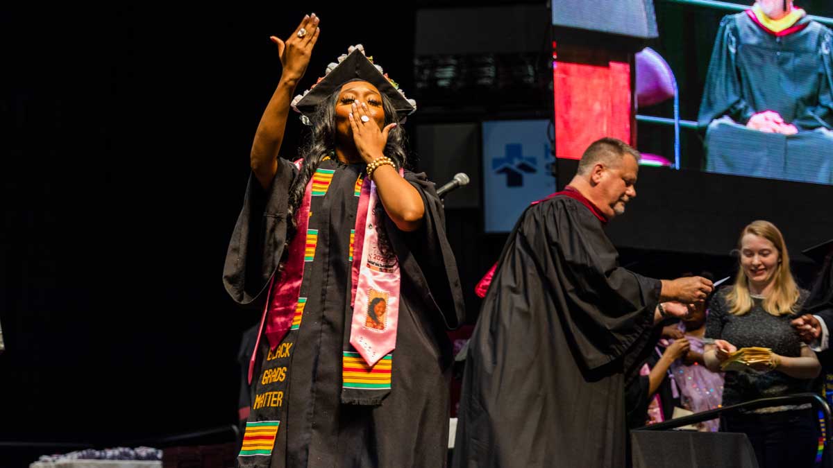 A black woman, dressed in commencement regalia, is seen blowing kisses to the crowd as she graduates.